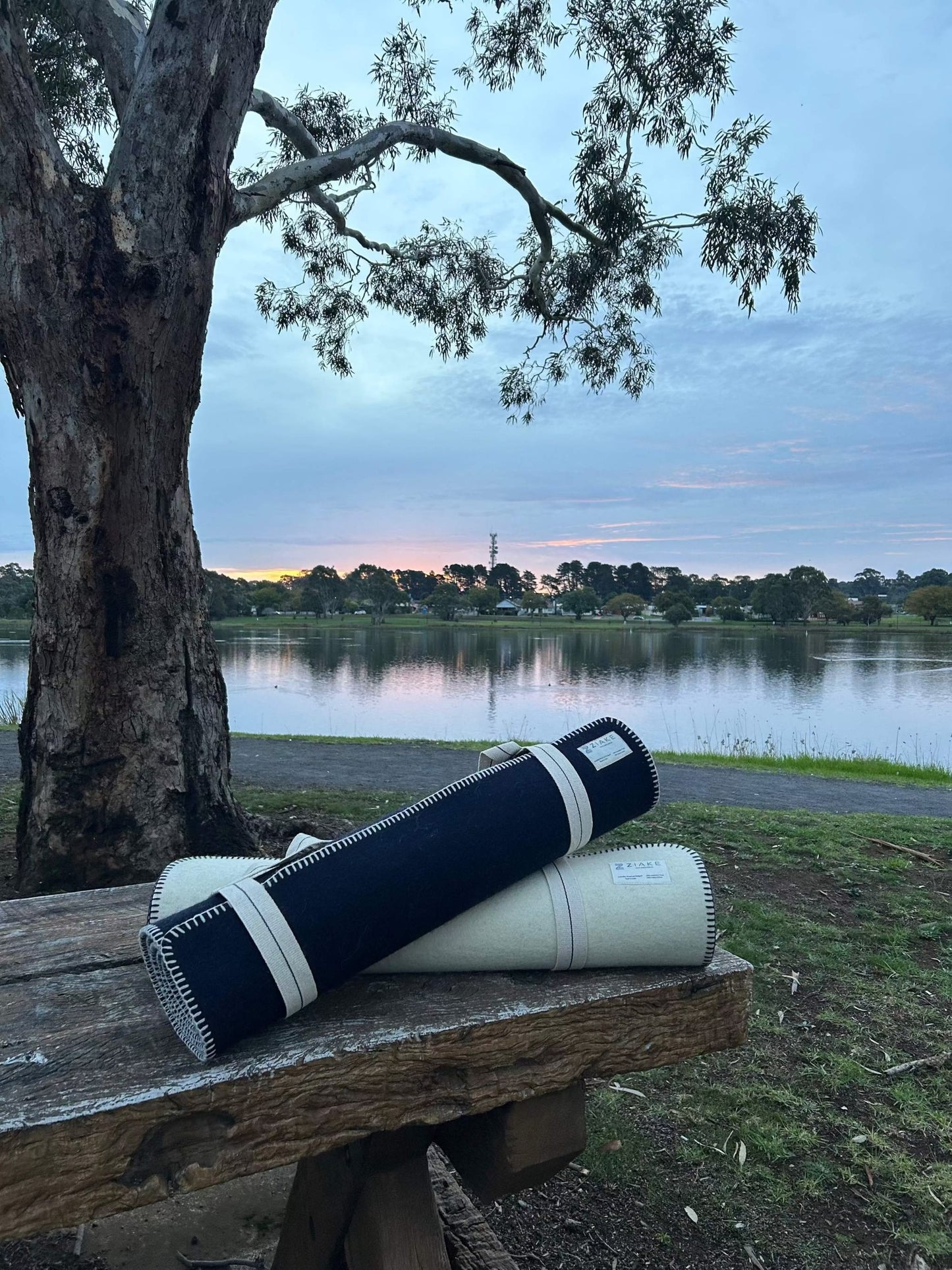 Australian crossbred wool fitness yoga mats in natural wool colour and navy colour, with cotton stitching and carry handle for yoga, pilates, meditation, fitness warm down and stretching.  In a sheep paddock with a lake in the background on sunset