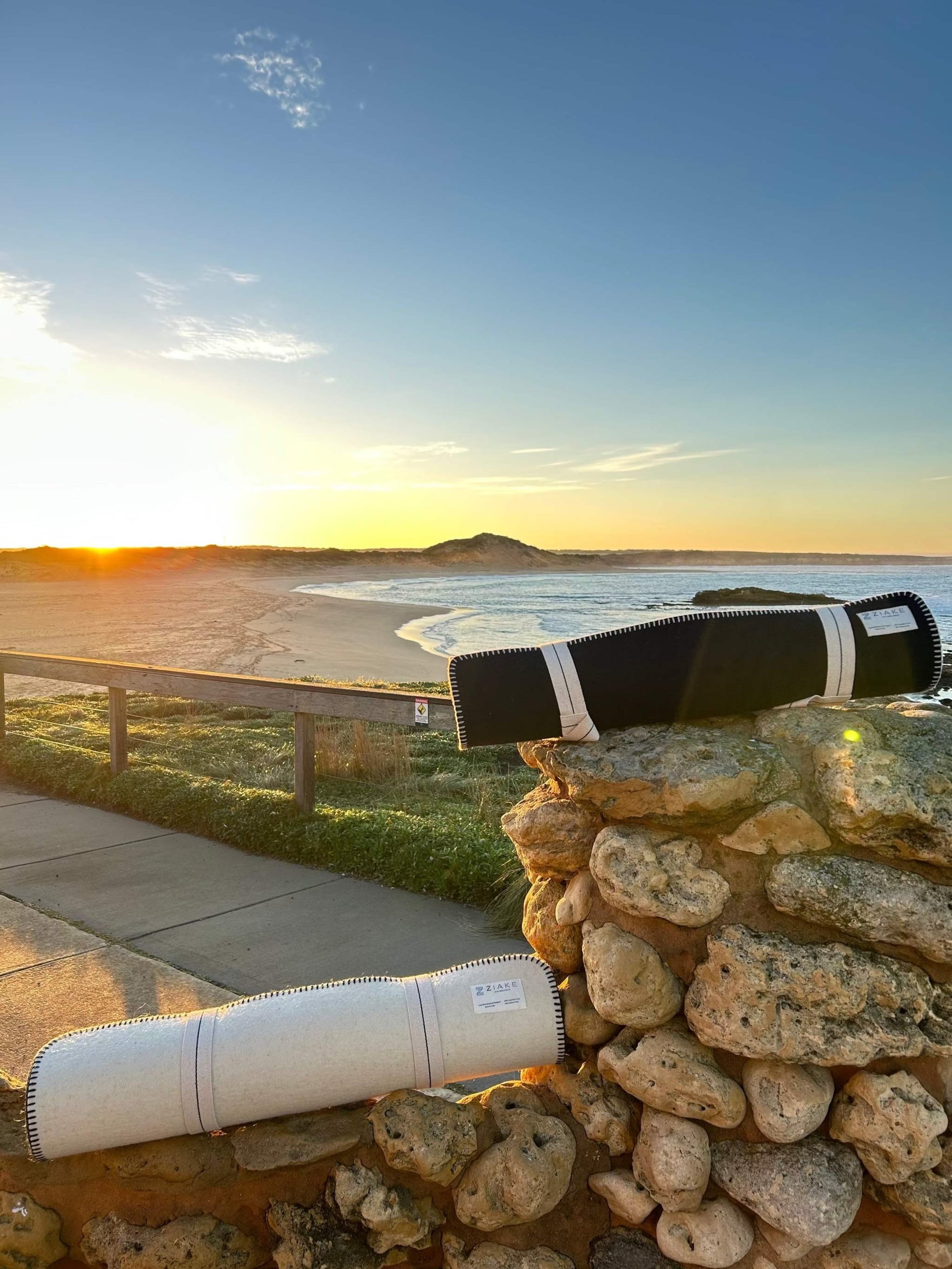 Australian crossbred wool fitness yoga mats in natural wool colour and navy colour, with cotton stitching and carry handle for yoga, pilates, meditation, fitness warm down and stretching. mats sitting on a stone wall in front of the beach, on sunrise. 